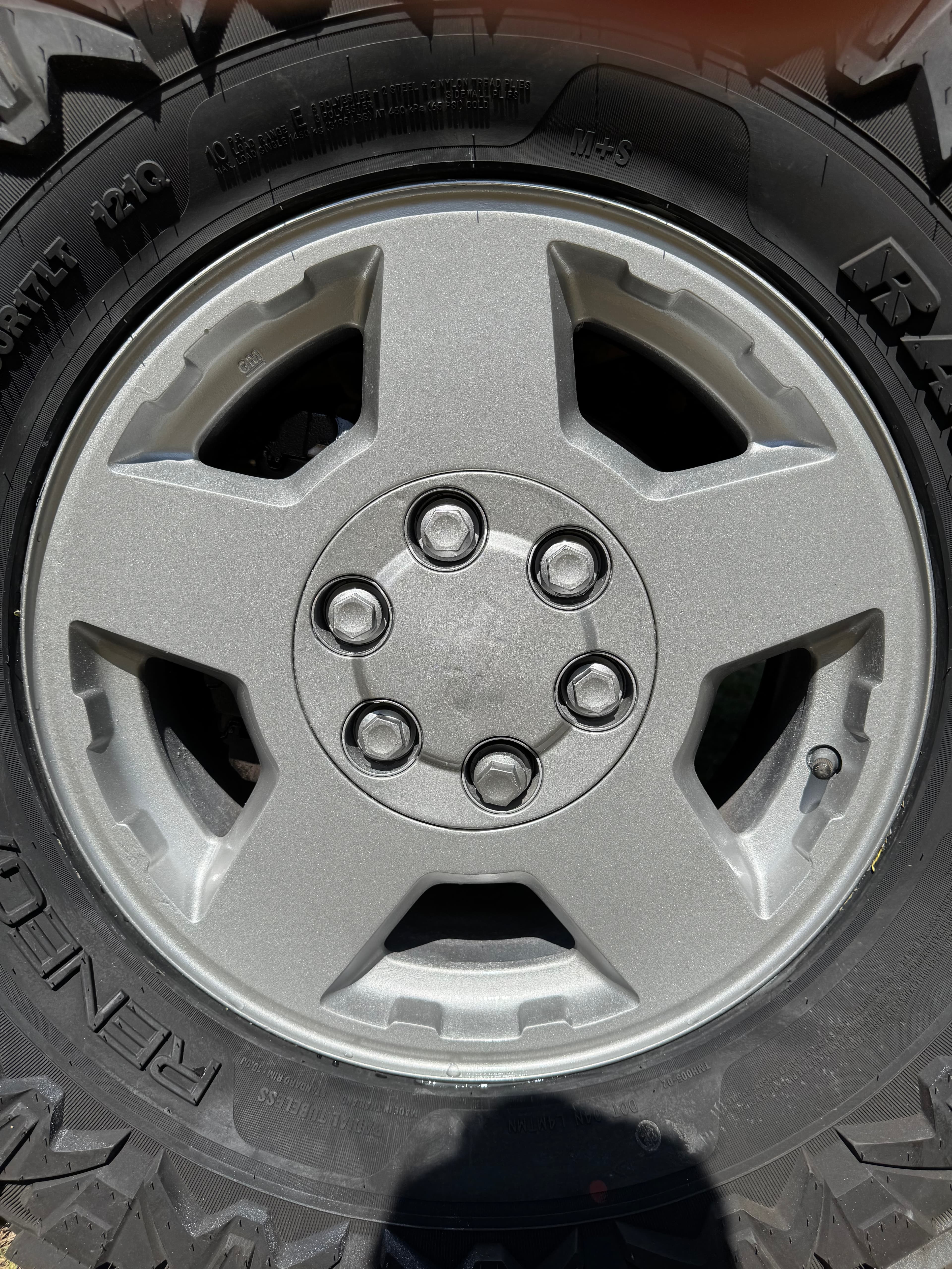 Silver five-spoke Chevrolet wheel rim with six lug nuts and a black off-road tire.