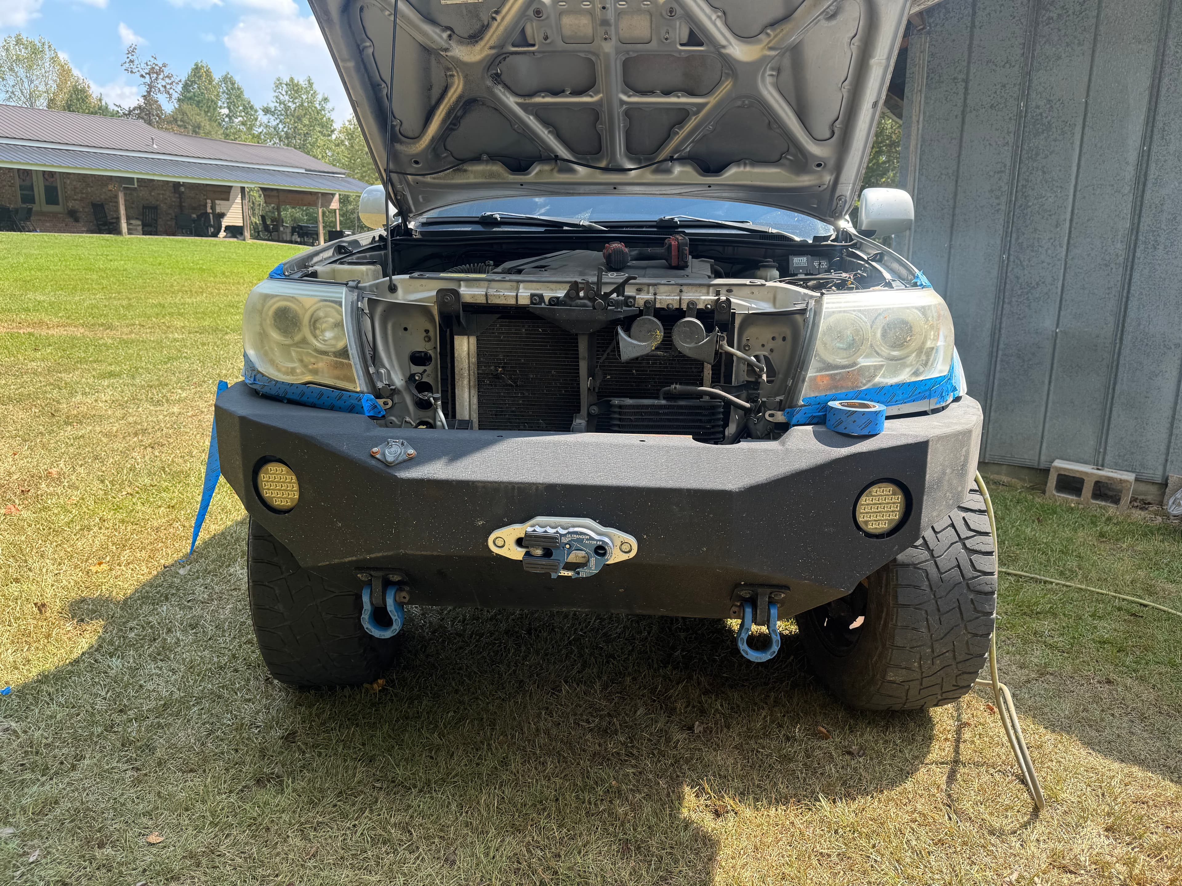 Silver truck with open hood and black aftermarket bumper, blue painter's tape on headlights.