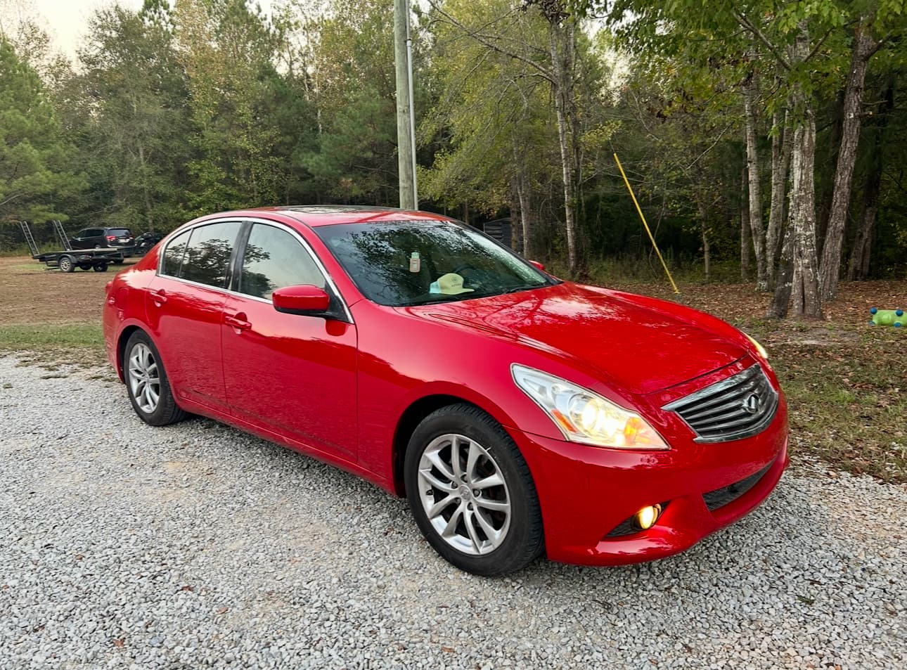 A bright red Infiniti sedan parked on a gravel driveway with its headlights turned on.