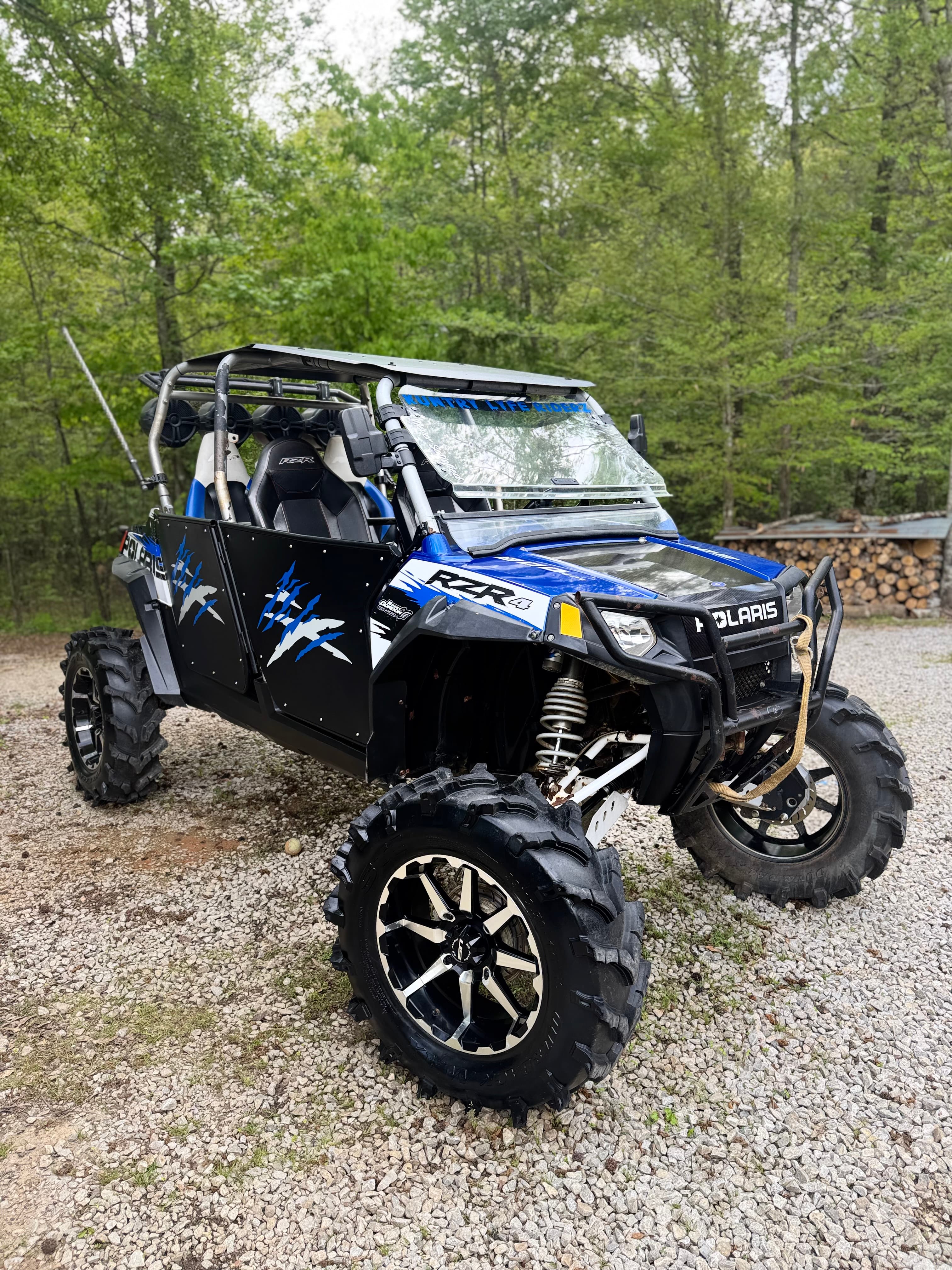 Blue and black Polaris RZR 4 off-road vehicle with large mud tires parked on gravel.