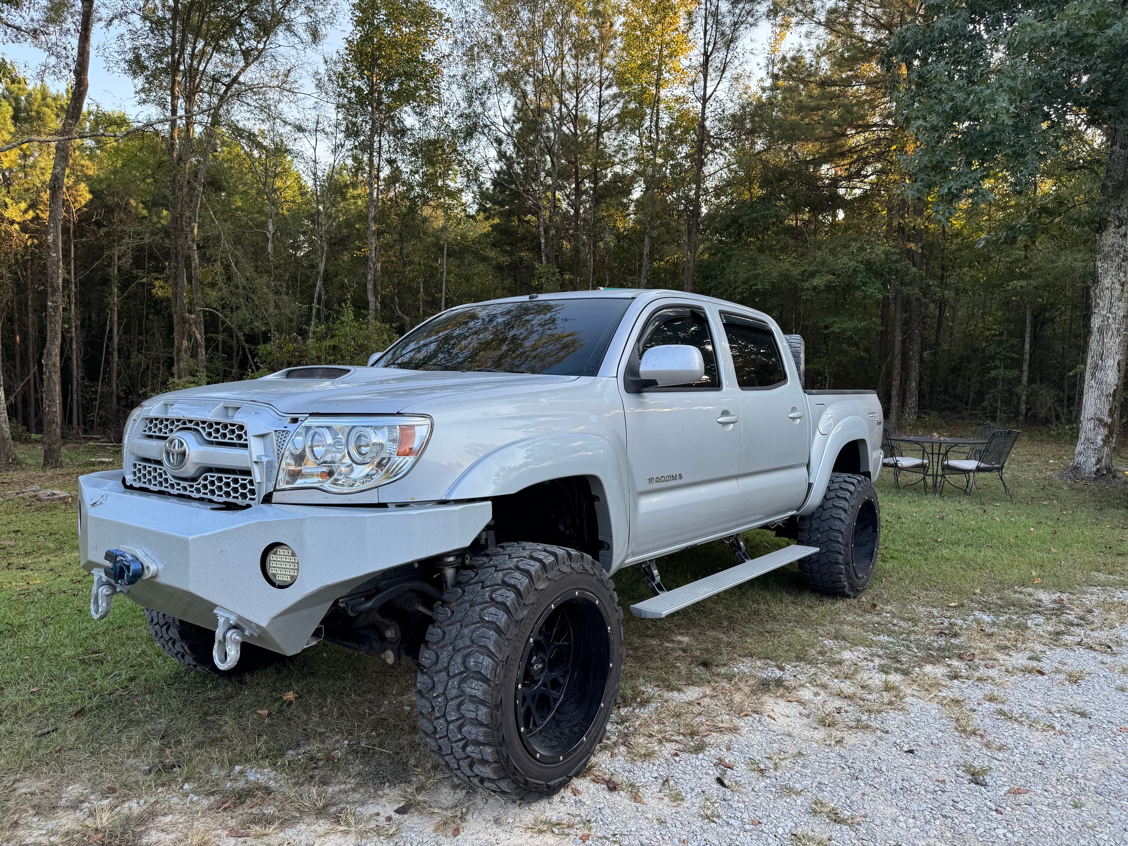 Lifted silver Toyota Tacoma with oversized off-road tires parked on gravel near a forest.