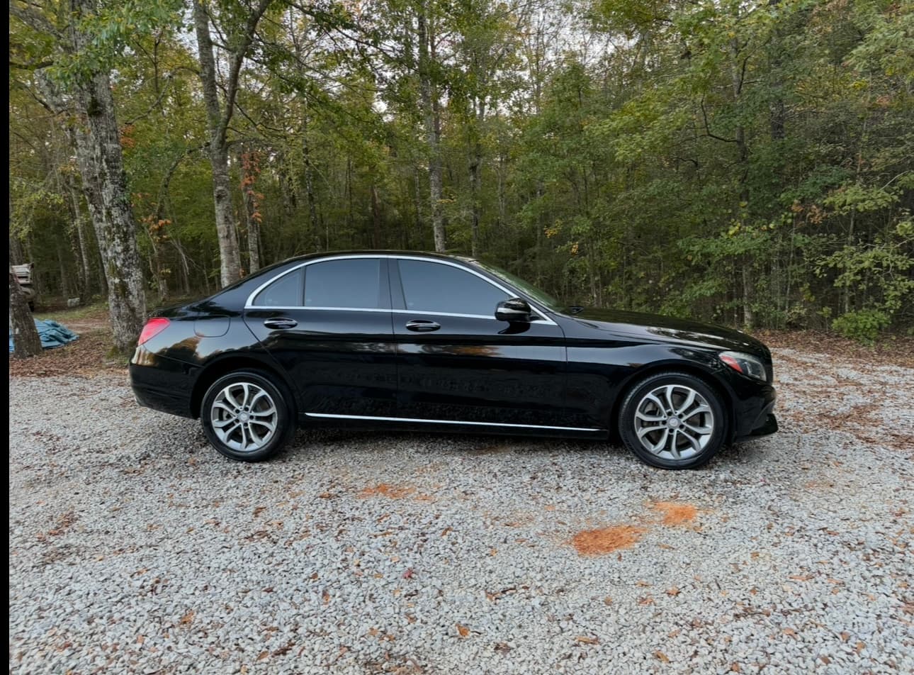 Black Mercedes-Benz sedan with tinted windows parked on a gravel driveway against dense green trees.